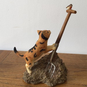 Puede incluir: Figura de un gato atigrado naranja con rayas negras, trepando por una horca de madera. Un pequeño pájaro se posa en la parte superior de la horca. El gato está sobre una base marrón texturizada. La figura está sobre una superficie de madera.