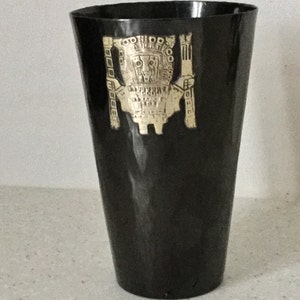 May include: Black drinking glass with a gold-coloured embossed design of a figure. The glass has a slightly flared shape. The embossed design is centered on the glass. The glass is likely made of a durable material.
