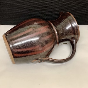 Może przedstawiać: Ręcznie robiony ceramiczny dzbanek z ciemnobrązowym szkliwem i czerwonobrązowymi i szarymi akcentami. Dzbanek ma zakrzywiony uchwyt i dzióbek do nalewania. Ceramika jest ustawiona pod kątem, pokazując podstawę i wnętrze.