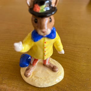 以下が含まれることがあります： 黒い帽子と花のアクセント、黄色のジャケット、青い蝶ネクタイを着たネズミの陶器製フィギュア。 ネズミは青いバケツを持っており、クリーム色の台座の上に立っています。 フィギュアは茶色と白のストライプの脚をしています。