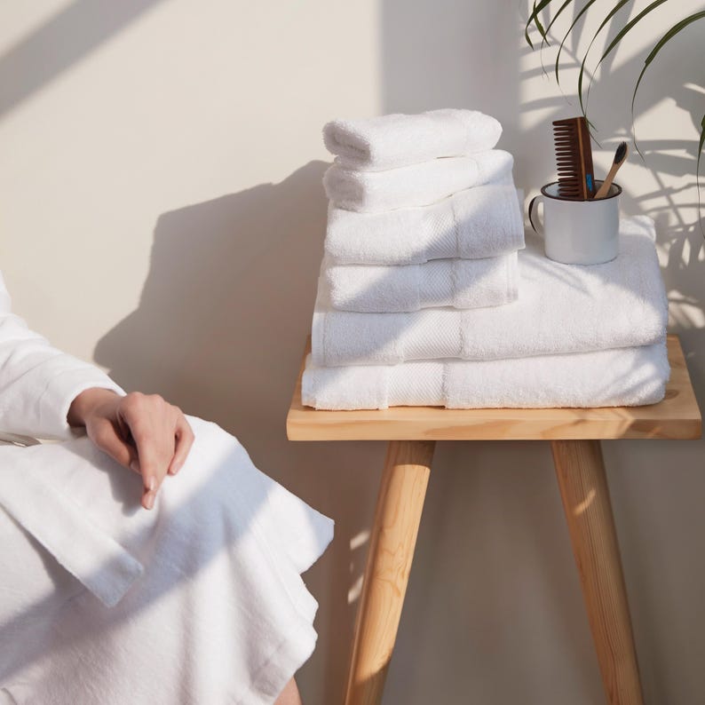 May include: A stack of white bath towels on a small wooden stool. The towels are neatly folded and arranged, with varying sizes visible. A white mug with a comb and toothbrush sits nearby. The scene is bathed in natural light, creating soft shadows.
