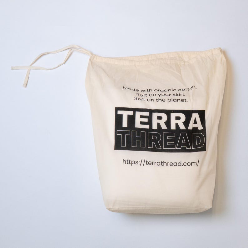 May include: A cream-colored drawstring bag with the text "TERRA THREAD" in a black rectangular box. The bag also features the text "Made with organic cotton. Soft on your skin. Soft on the planet." and a website address.