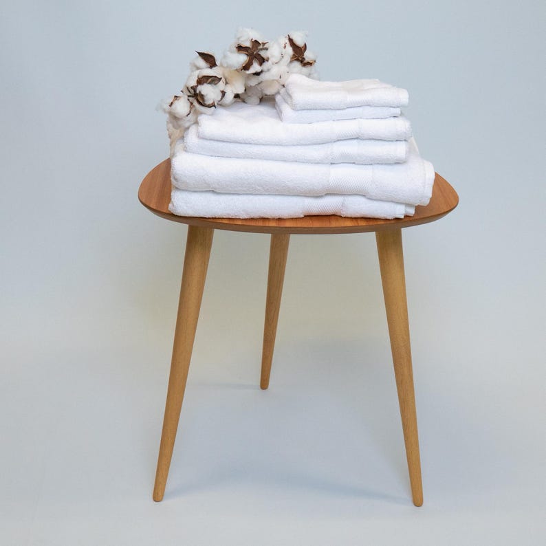 May include: A stack of folded white towels on a small, round, wooden side table with three angled legs. Cotton sprigs with brown and white accents are arranged on top of the towels. The table is set against a light blue background.