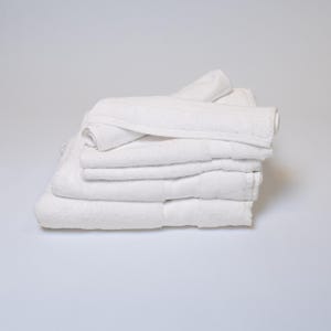 May include: A stack of clean, white bath towels. The towels are neatly folded and stacked, with a rolled-up towel on top. The towels are made of a soft, absorbent material.