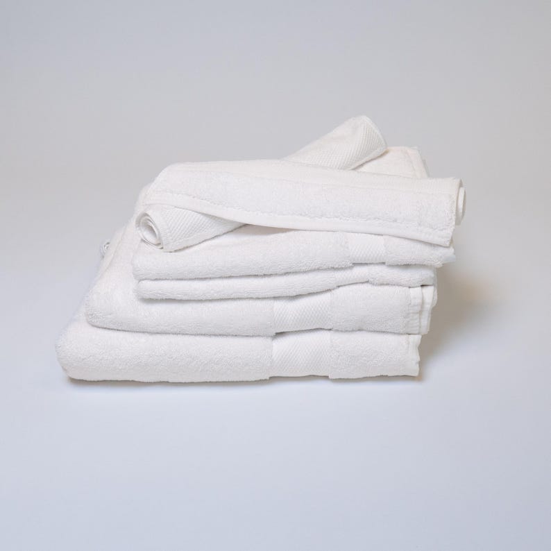 May include: A stack of clean, white bath towels. The towels are neatly folded and stacked, with a rolled-up towel on top. The towels are made of a soft, absorbent material.