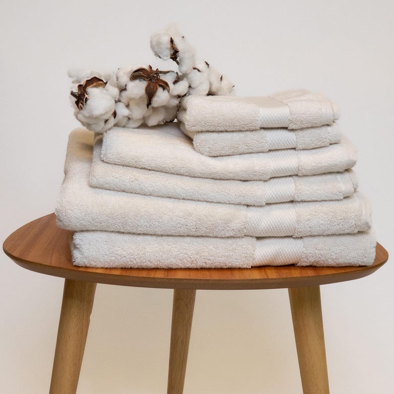 May include: A stack of cream-colored bath towels of various sizes, neatly arranged on a small, round wooden table with three legs. A sprig of cotton bolls rests on top of the towels. The towels have a textured appearance.
