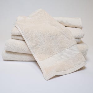 May include: A stack of beige bath towels. The towels are folded and stacked on top of each other. The top towel has a textured border. The towels are made of a soft, absorbent material.