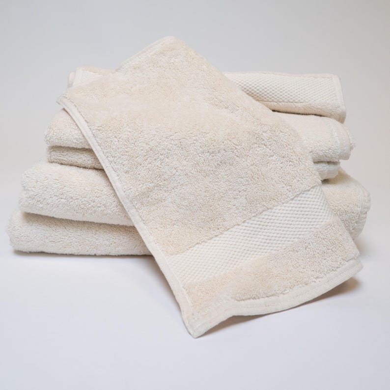 May include: A stack of beige bath towels. The towels are folded and stacked on top of each other. The top towel has a textured border. The towels are made of a soft, absorbent material.