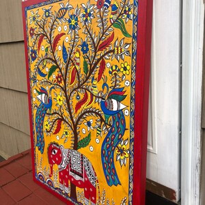Puede incluir: Una pintura colorida sobre lienzo que representa un &aacute;rbol con pavos reales, p&aacute;jaros y un elefante. El &aacute;rbol est&aacute; pintado en tonos de amarillo, verde y marr&oacute;n, con flores en rojo, azul y blanco. Los pavos reales son azules y verdes con colas largas. El elefante es rojo con detalles blancos. La pintura est&aacute; enmarcada en un borde rojo.