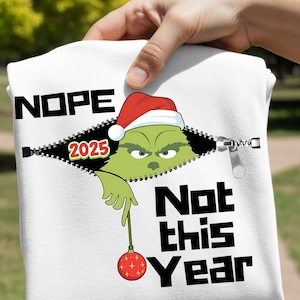 Könnte beinhalten: Weißes T-Shirt mit einem Grinch-Motiv, das einen Weihnachtsmannhut trägt und aus einem Reißverschluss-Design herausschaut. Der Text "NOPE 2025" steht über dem Reißverschluss und "Not this Year" darunter. Der Grinch hält eine rote Christbaumkugel.