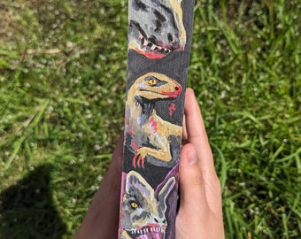 May include: A wooden block painted with three different dinosaur heads. The top dinosaur is gray with a black mouth and red teeth. The middle dinosaur is yellow with a black mouth and red teeth. The bottom dinosaur is purple with a black mouth and red teeth.