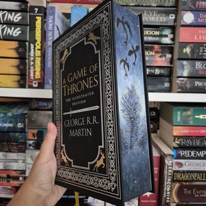 Könnte beinhalten: Ein gebundenes Exemplar von "A Game of Thrones: The Illustrated Edition" von George R.R. Martin. Das Buch hat einen schwarzen Einband mit goldenen Akzenten, Hirsch- und Drachenillustrationen und einen blauen Buchrücken mit einem Baumdesign. Das Buch wird vor einem Bücherregal gehalten.