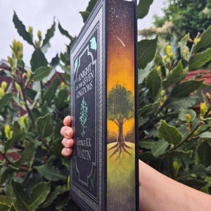 Op de afbeelding: Een hardcover boek getiteld "A Knight of the Seven Kingdoms" van George R.R. Martin. Het boek heeft een zwarte kaft met zilveren accenten en een geverfde zijkant met een boom en een zonsondergang. Het boek wordt tegen een achtergrond van groen gebladerte gehouden.