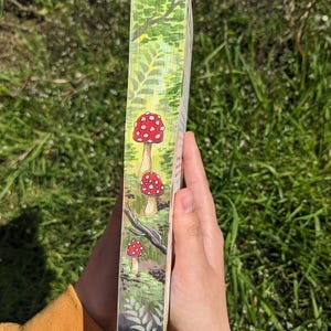 May include: A painted wooden book spine with a green forest scene and three red and white mushrooms. The mushrooms are painted in a whimsical style with white spots.