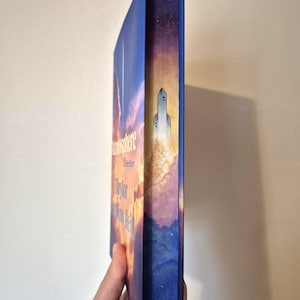 Op de afbeelding: Een hardcover boek getiteld "Atmosphere: A Novel" van Taylor Jenkins Reid. De rug van het boek toont een blauwe en paarse lucht met een raketillustratie. De omslag toont een zonsondergangscène met de titel in gouden letters.