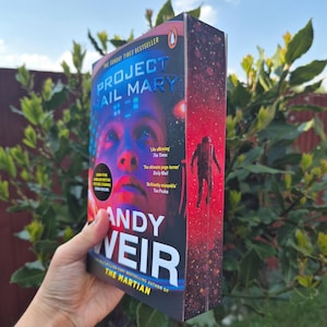 May include: A copy of "Project Hail Mary" by Andy Weir, a Sunday Times bestseller. The book's cover displays a person's face in blue and purple tones, with the title in white and blue. The spine shows an astronaut against a red backdrop. The book is held up.