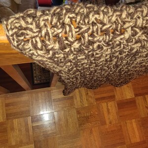 May include: A close-up of a brown and beige crocheted blanket. The blanket has a textured, woven appearance with a scalloped edge. The color palette is neutral, with a mix of light and dark brown yarns. The blanket is draped over a wooden surface.