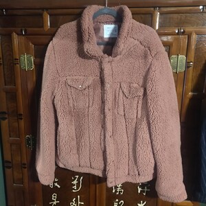 May include: A dusty rose-colored, button-down jacket with a textured, fuzzy exterior. The jacket features two chest pockets and a collared neckline. It is hanging on a black hanger against a dark wooden cabinet.