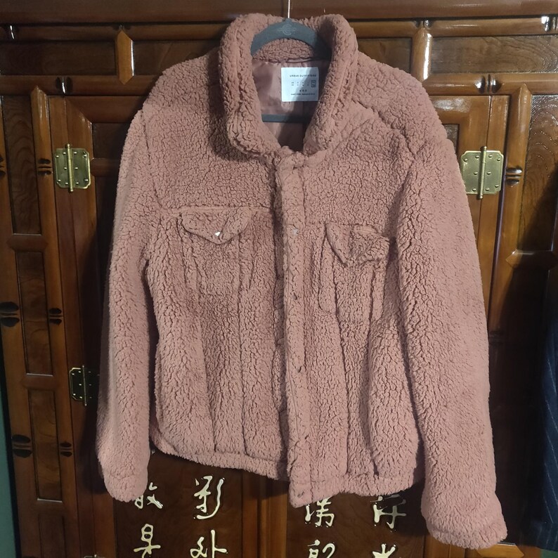 May include: A dusty rose-colored, button-down jacket with a textured, fuzzy exterior. The jacket features two chest pockets and a collared neckline. It is hanging on a black hanger against a dark wooden cabinet.