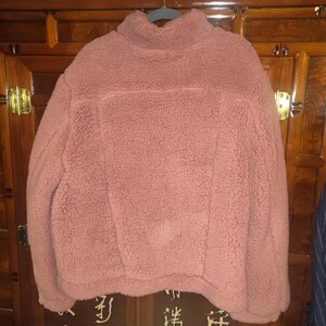 May include: A dusty rose-colored fleece pullover with a high collar. The soft, textured fabric is visible, and the garment is hanging on a gray hanger. The pullover has a simple, classic design, suitable for casual wear.