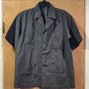 May include: A dark grey short-sleeved button-up shirt with multiple pockets and a collar. The shirt has a textured fabric and a relaxed fit.