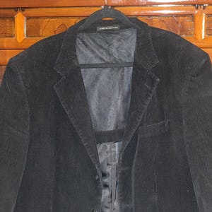 以下が含まれることがあります： ノッチドラペル、2つのフロントポケット、2つボタン留めの黒いコーデュロイブレザー。ブレザーは黒いハンガーにかかっています。内側の裏地が見えます。「croft & barrow」のブランド名がハンガーに印刷されています。