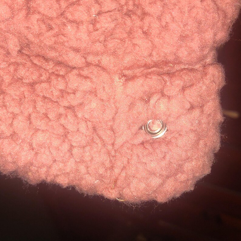 May include: Close-up of a section of a dusty rose-colored, textured fabric. A silver-colored, circular metal snap is visible on the fabric. The fabric appears to be a soft, fuzzy material.