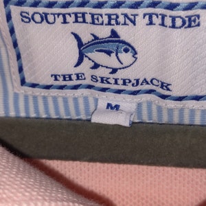 May include: A white tag with blue trim and the text "Southern Tide The Skipjack" and a blue and white fish illustration. The tag has a smaller white tag attached with the letter "M" printed on it.