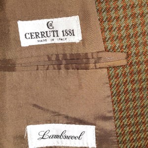 以下が含まれることがあります： ツイードジャケットの内側のクローズアップ。茶色の裏地には「CERRUTI 1881 MADE IN ITALY」と書かれたラベルが付いています。別のラベルには「Lambswool」と書かれています。ツイード生地は緑とオレンジの混合です。
