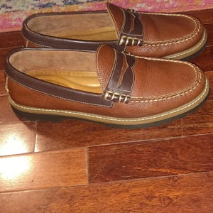 May include: A pair of brown leather penny loafers. The shoes have a classic design with a rounded toe, a strap across the vamp, and a stitched sole. The leather is a rich brown color with darker brown accents.