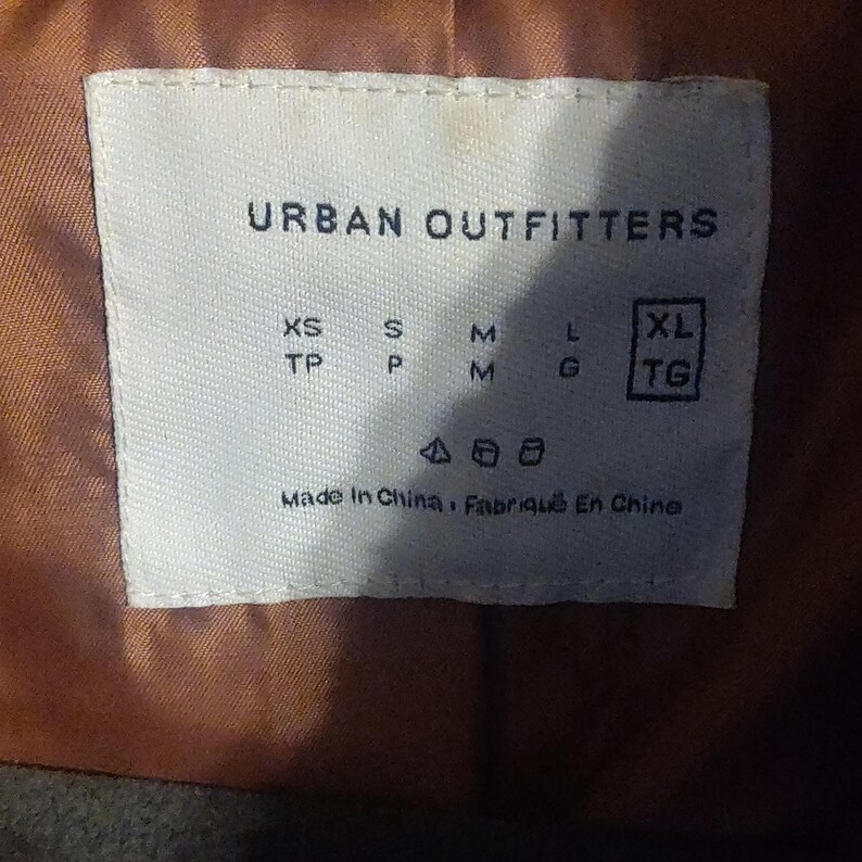 May include: A white label with the text "URBAN OUTFITTERS" and size indicators: XS, S, M, L, and XL. The label also includes the text "Made In China, Fabriqu&eacute; En Chine". The label is on a brown fabric.