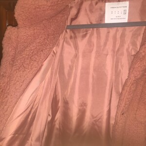May include: A dusty rose-colored teddy bear coat with a satin lining. The coat is open, revealing the interior lining and the texture of the outer material. The tag reads "Urban Outfitters".