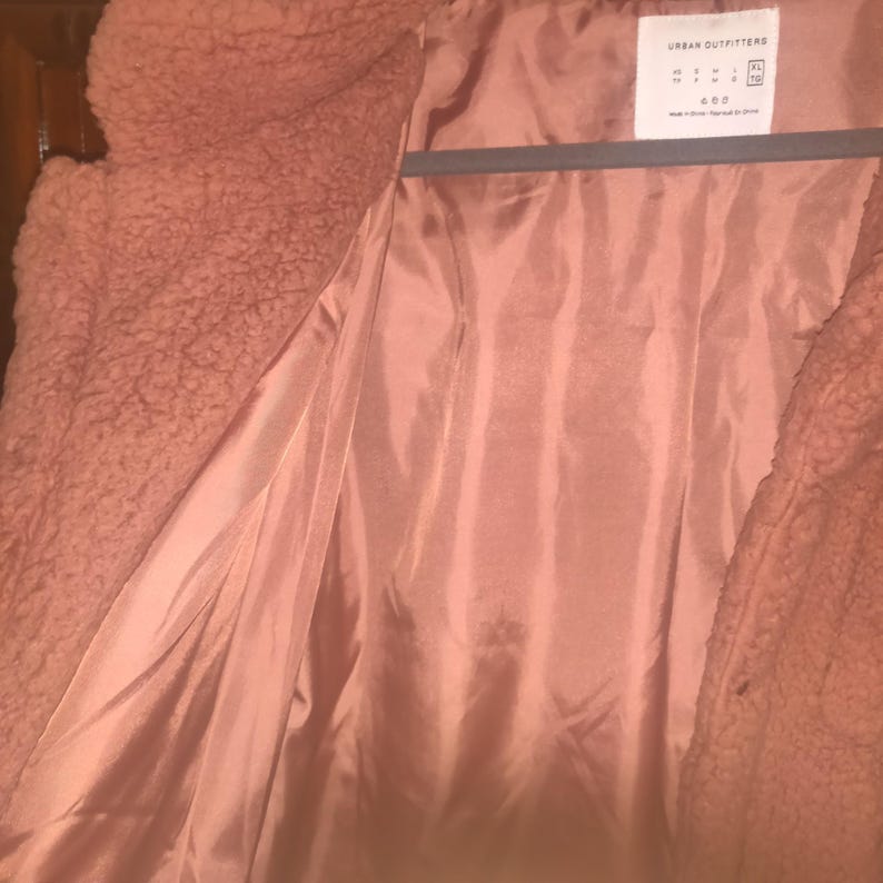 May include: A dusty rose-colored teddy bear coat with a satin lining. The coat is open, revealing the interior lining and the texture of the outer material. The tag reads "Urban Outfitters".