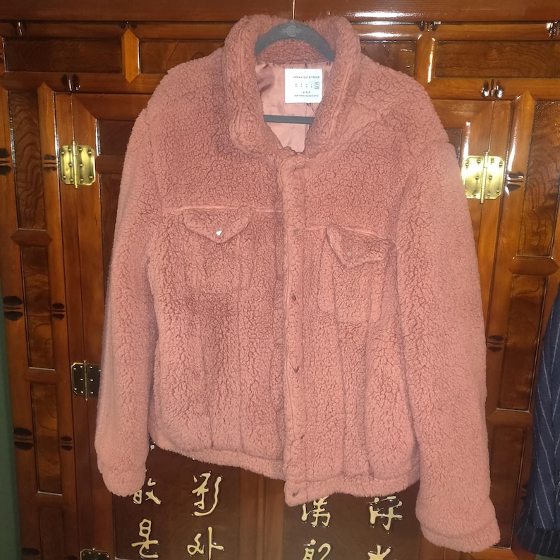 May include: A dusty rose-colored, plush jacket with a button-up front, two chest pockets, and a collared neckline. The jacket is made of a soft, textured material and hangs on a black hanger.