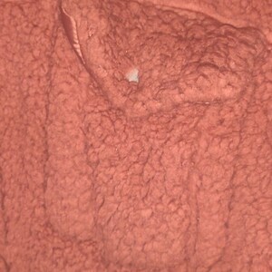May include: Close-up of a textured, reddish-brown fabric. The material has a dense, looped pile, creating a soft, plush appearance. The fabric's surface is uneven, with variations in texture and color. A small, light-colored area is visible.