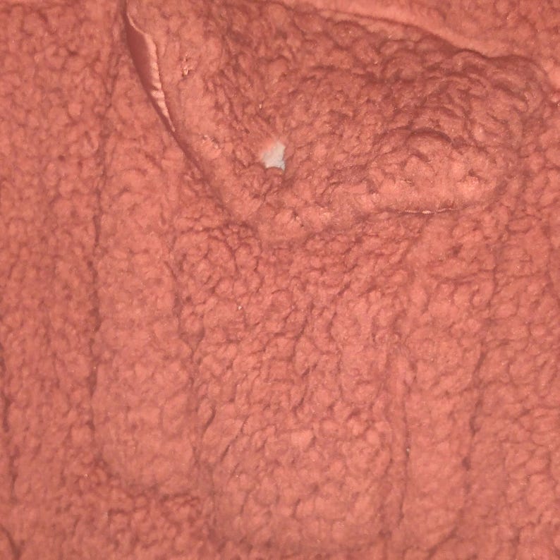 May include: Close-up of a textured, reddish-brown fabric. The material has a dense, looped pile, creating a soft, plush appearance. The fabric's surface is uneven, with variations in texture and color. A small, light-colored area is visible.