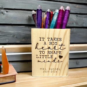 May include: A wooden pen holder with the engraved message "IT TAKES A BIG HEART TO SHAPE LITTLE MINDS" and "MRS. RUSSEL 2025-2026". The holder contains various colored gel pens. The holder is on a wooden desk.