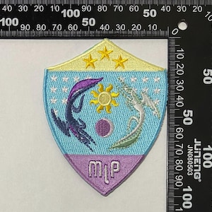 Op de afbeelding: Een blauwe en paarse geborduurde patch in de vorm van een schild. Het schild heeft een gele achtergrond met vijf sterren, een zon, een maan en twee gestileerde wezens. De tekst "MyP" is in paars geborduurd aan de onderkant van het schild.