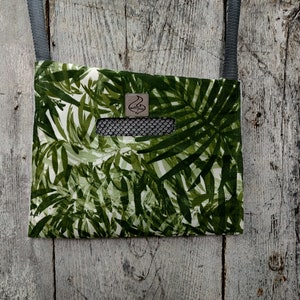 May include: A square crossbody bag with a green and white tropical leaf pattern. It features a black mesh pocket and a grey strap. A small, square leather patch with a logo is sewn on the bag.