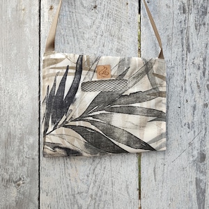 May include: A square crossbody bag featuring a beige and black leaf design. It has a brown strap and a small mesh pocket. A small leather tag is sewn on the front of the bag.