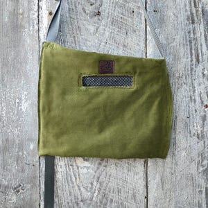 May include: Olive green corduroy shoulder bag. It has a rectangular shape, a mesh pocket, and a brown leather logo patch. The bag is suspended by a grey strap.