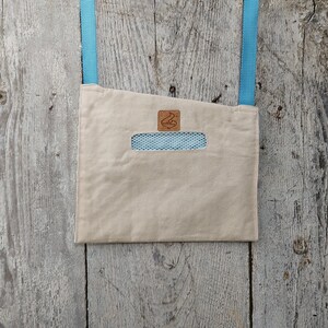 Op de afbeelding: Beige canvas crossbody tas met een rechthoekige uitsparing die een blauw mesh interieur onthult. De tas heeft een lichtblauwe band en een klein bruin leren logo patch. Op een verweerde houten achtergrond.