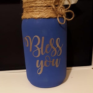 Op de afbeelding: Een blauwe geverfde glazen pot met een deksel van jute en een jutebloem. De pot heeft een zilveren vinyl sticker met de tekst "Bless you".