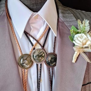 Puede incluir: Una blazer gris sobre una camisa blanca con cuello, adornada con tres corbatas bolo con colgantes circulares decorativos. Un ojal de rosa blanca con acentos de vegetación y cinta adorna la solapa. Las corbatas bolo tienen cordones de cuero trenzado.