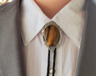 May include: A silver bolo tie with a brown and gold tiger's eye stone set in a decorative frame. The bolo tie is worn on a white shirt with a gray suit jacket.