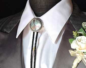 May include: A black bolo tie with a silver and abalone shell bolo tie slide. The bolo tie is worn on a white dress shirt with a gray suit jacket. A white flower boutonniere is pinned to the jacket.
