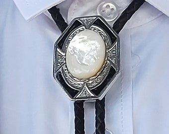 Puede incluir: Un bolo tie plateado con un colgante ovalado con un patrón en blanco y negro que presenta una piedra blanca similar a una perla. La corbata está hecha de cordón trenzado negro.