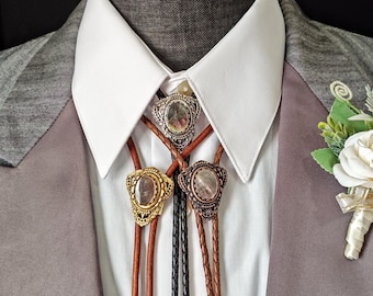 May include: Three bolo ties with decorative metal slides and leather cords are displayed against a white collared shirt and gray blazer. The slides feature ornate designs and gemstones. One is gold, one is silver, and one is bronze.