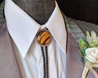 May include: A brown and gold bolo tie with a tiger's eye stone. The bolo tie is worn with a white shirt and a gray suit jacket.