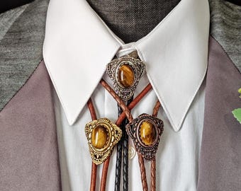 May include: Three bolo ties with decorative metal slides and oval tiger's eye stones. The bolo ties have brown leather cords and a black braided cord. The ties are displayed on a white collared shirt and a gray blazer.